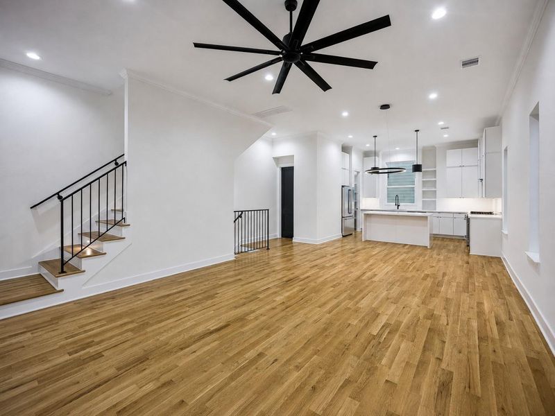 The open main living level offers an impressive sense of space with warm wood flooring, recessed lighting, crown molding, and an oversized contemporary ceiling fan. The modern kitchen sits just beyond, creating an easy connection between cooking, dining, and gathering areas. A sculptural staircase with black iron rails adds architectural character while maintaining the home’s clean, sophisticated style. The open main living level offers an impressive sense of space with warm wood flooring, recessed lighting, crown molding, and an oversized contemporary ceiling fan. The modern kitchen sits just beyond, creating an easy connection between cooking, dining, and gathering areas. A sculptural staircase with black iron rails adds architectural character while maintaining the home’s clean, sophisticated style.