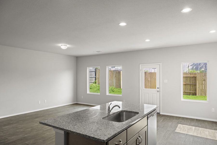 A kitchen and living room with a grey marble countertop island, vinyl flooring, white painted walls, white-framed window, and a white door.