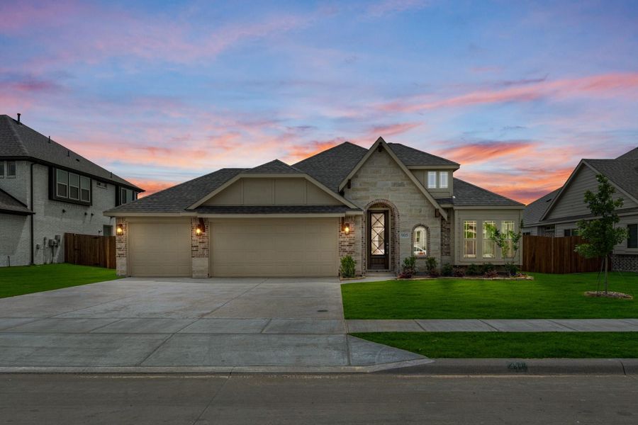 Single-story home with stone and brick exterior, three-car garage, arched entry, and green front lawn at dusk Single-story home with stone and brick exterior, three-car garage, arched entry, and green front lawn at dusk