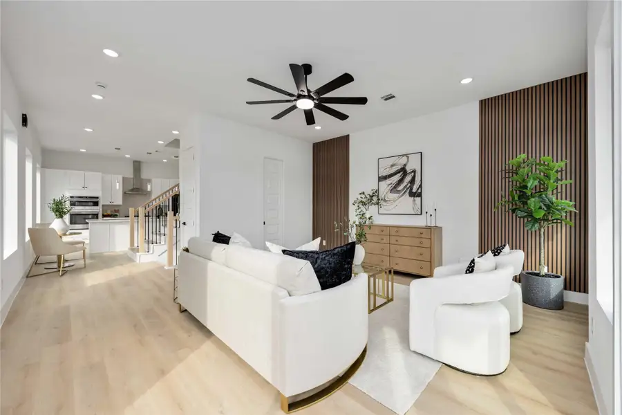 A bright, open living area seamlessly connects to the kitchen, featuring modern white furniture, wood-slat accent walls, and large windows that fill the space with natural light.