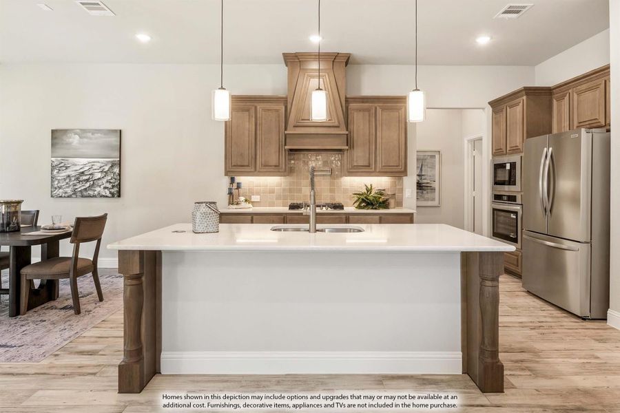 Kitchen with large white island, wood cabinets, custom range hood, and stainless steel appliances on light hardwood floors Kitchen with large white island, wood cabinets, custom range hood, and stainless steel appliances on light hardwood floors
