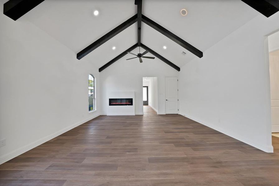 Unfurnished living room featuring beam ceiling, dark wood finished floors, a glass covered fireplace, ceiling fan, and high vaulted ceiling