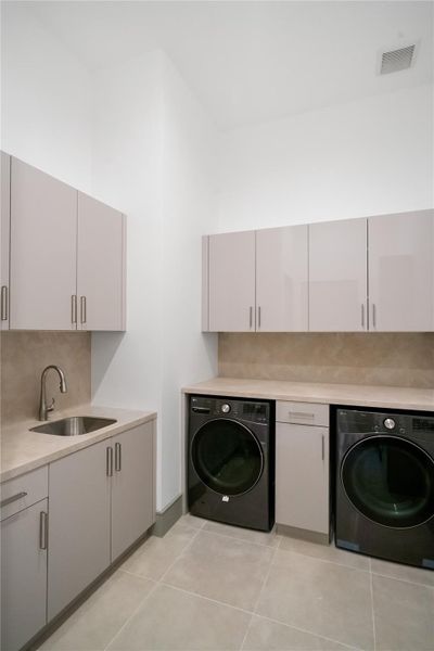 This photo shows a modern laundry room featuring sleek cabinetry, a built-in sink, and a washer and dryer set. The space is designed with neutral tones and ample storage, offering a functional and stylish utility area.