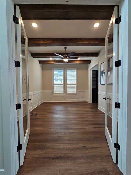This photo showcases a room with elegant French doors, rich wood flooring, and exposed wooden ceiling beams. The space has a ceiling fan, recessed lighting, and a window with shutters, creating a bright, inviting atmosphere ideal for an office or study.