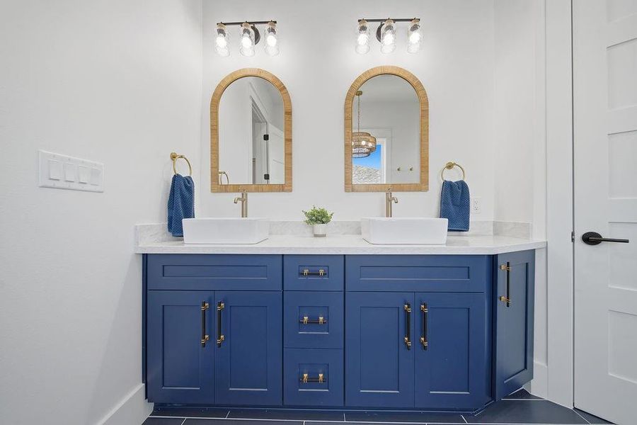 This bathroom features a stylish double vanity with bold blue cabinetry and gold hardware. It includes two vessel sinks, arched mirrors, and modern lighting, creating a bright and elegant space.