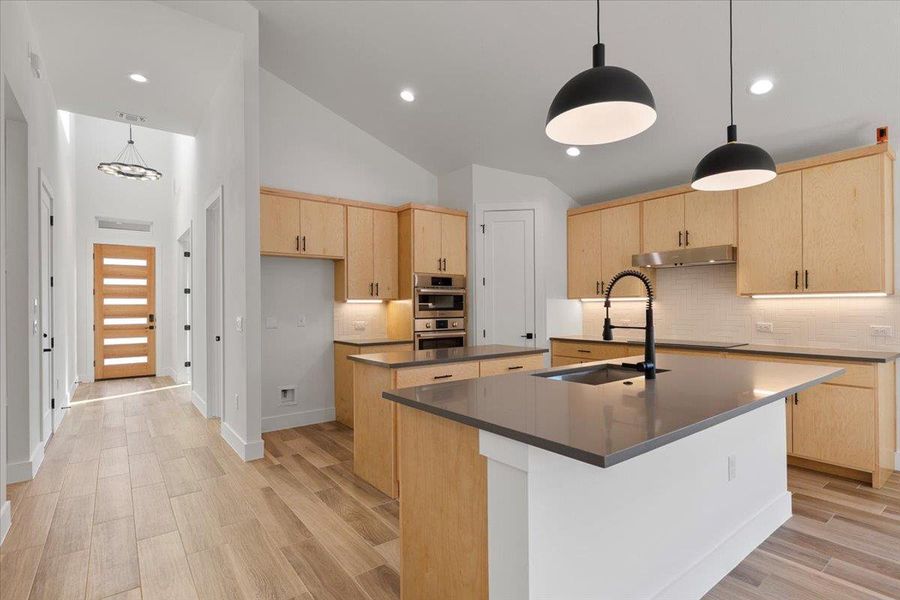 Kitchen featuring light brown cabinetry, backsplash, decorative light fixtures, a center island with sink, and wood tiled floors Kitchen featuring light brown cabinetry, backsplash, decorative light fixtures, a center island with sink, and wood tiled floors
