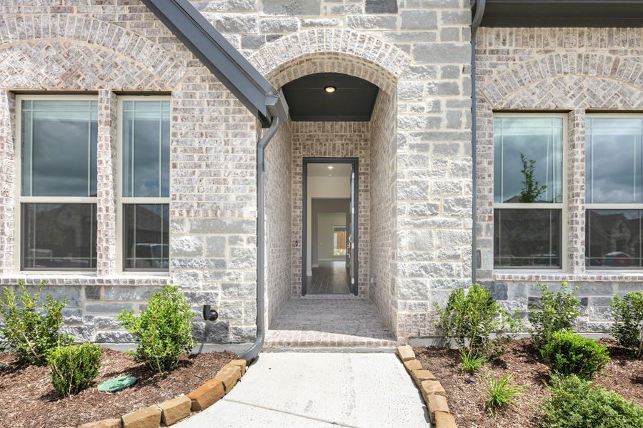 Home entrance with arched brick surround, dark front door, concrete walkway, and small landscaped shrub beds on each side. Home entrance with arched brick surround, dark front door, concrete walkway, and small landscaped shrub beds on each side.