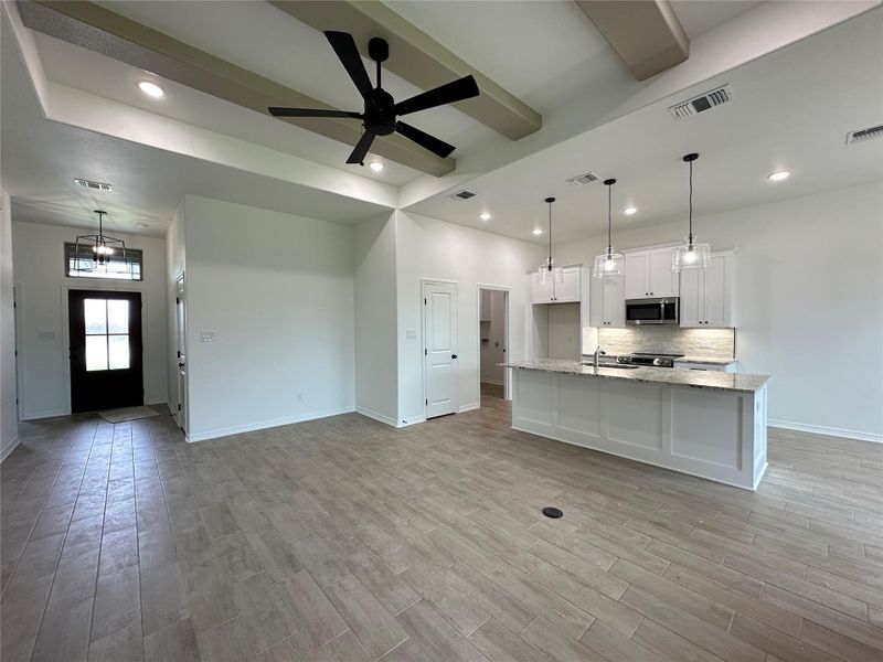 Spacious open-concept interior featuring light wood-finish flooring, a contemporary kitchen island with pendant lighting, recessed ceiling lights, and exposed ceiling beams