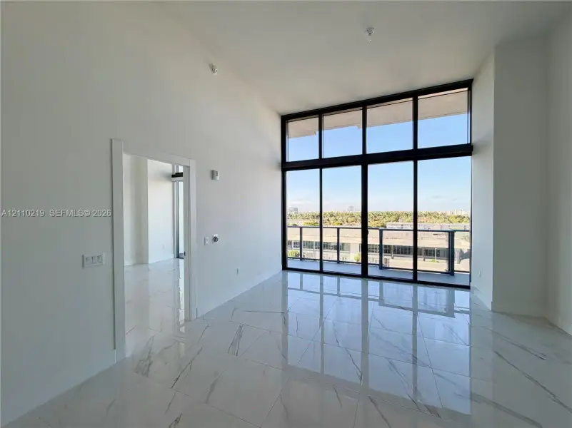 High-ceiling interior featuring polished stone-finish floor tiling, expansive floor-to-ceiling windows with black metal framing, and a balcony with a glass and metal railing