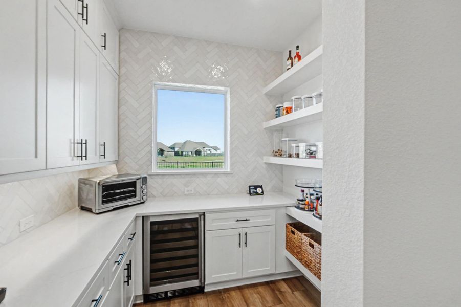 A well-appointed butler's pantry or coffee station includes custom cabinetry, additional white countertops, and a full-height tile backsplash. Open shelving displays glassware and barware, while a dedicated wine refrigerator and built-in toaster oven add convenience.