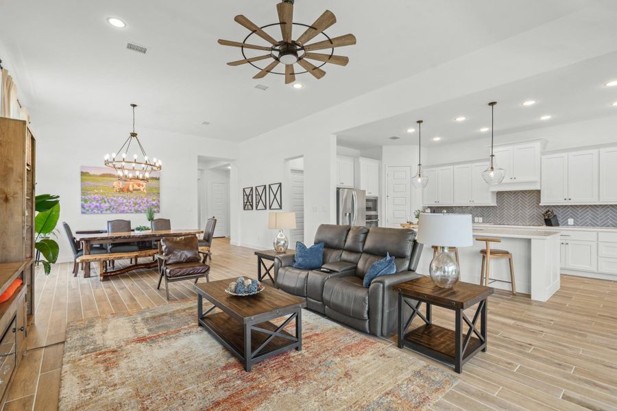 This spacious open-concept living area features a modern kitchen with white cabinetry, pendant lighting, and a stylish herringbone backsplash. The room is bright and airy, with wood-look tile flooring, a cozy sitting area, and a dining space illuminated by a chic chandelier. Ideal for entertaining and family gatherings.