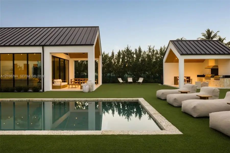 Outdoor living area featuring a rectangular pool with stone coping, artificial turf lawn, covered patio, and built-in outdoor kitchen with metal roof structures