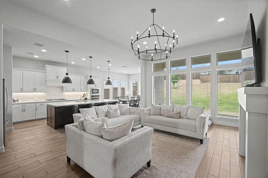 Open-concept living space featuring wood-finish flooring, a large island kitchen with white cabinetry and dark pendant lighting, and an expansive window wall overlooking the backyard