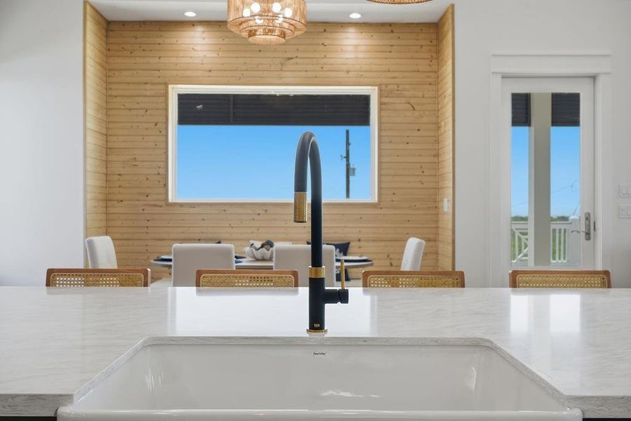 Modern kitchen with a sleek black faucet and white marble countertop. Features a wood-accented wall, large window, and natural light. Dining area in the background with elegant decor.