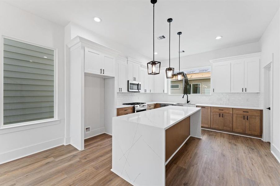 This modern kitchen features a spacious island with a sleek white countertop, pendant lighting, and a mix of white and wood cabinetry. The open layout is complemented by stainless steel appliances and light wood flooring, creating a bright and inviting space.