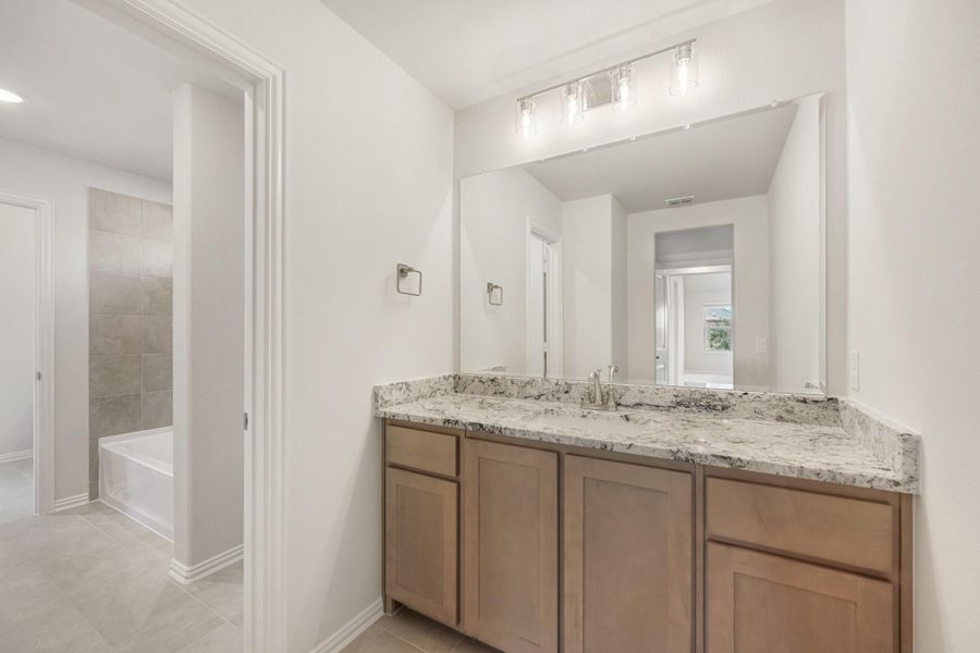 Bathroom with wood vanity, granite countertop, large mirror, and separate soaking tub with tile surround visible through doorway.