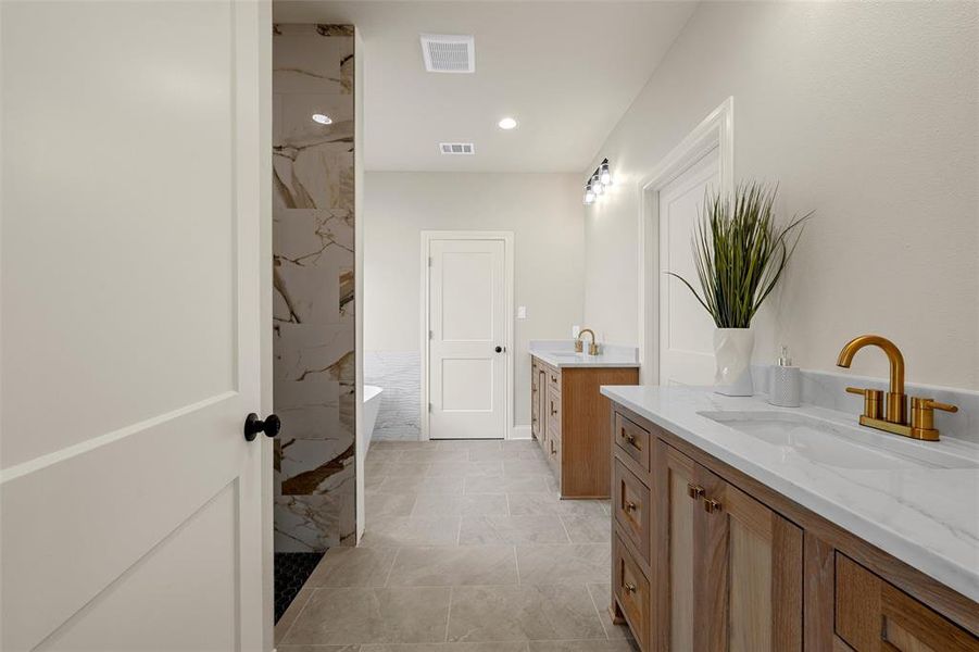Bathroom featuring dual wood-finish vanities with white countertops, gold-finish fixtures, a freestanding bathtub, and a walk-in shower with natural stone-look tiling