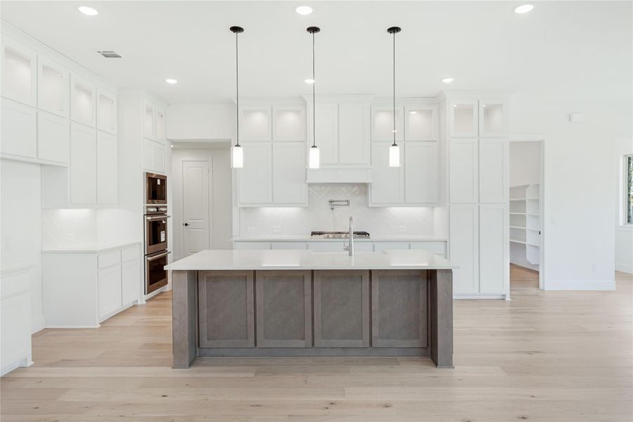 White kitchen with large gray island, pendant lights, double wall ovens, and light hardwood floors