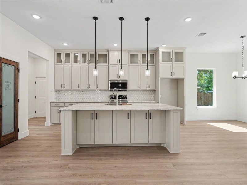 Kitchen featuring an expansive island with a built-in sink, stone countertop, pendant lighting, shaker-style cabinetry, and wood-finish flooring Kitchen featuring an expansive island with a built-in sink, stone countertop, pendant lighting, shaker-style cabinetry, and wood-finish flooring
