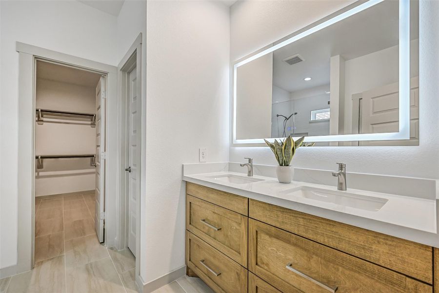 The primary bath features a long dual-sink vanity with warm wood-tone cabinetry, sleek fixtures and an impressive illuminated mirror, while an adjoining walk-in closet keeps storage conveniently close at hand. The primary bath features a long dual-sink vanity with warm wood-tone cabinetry, sleek fixtures and an impressive illuminated mirror, while an adjoining walk-in closet keeps storage conveniently close at hand.