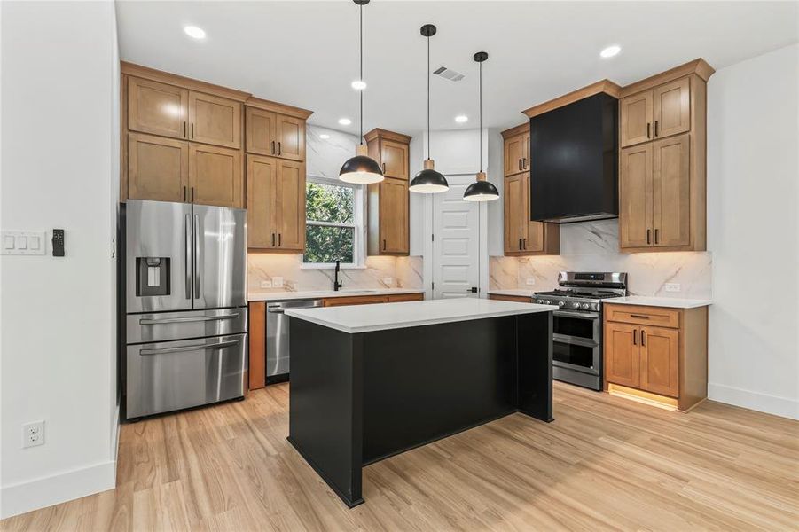 Kitchen featuring stainless steel appliances, decorative light fixtures, brown cabinetry, a center island, and ventilation hood