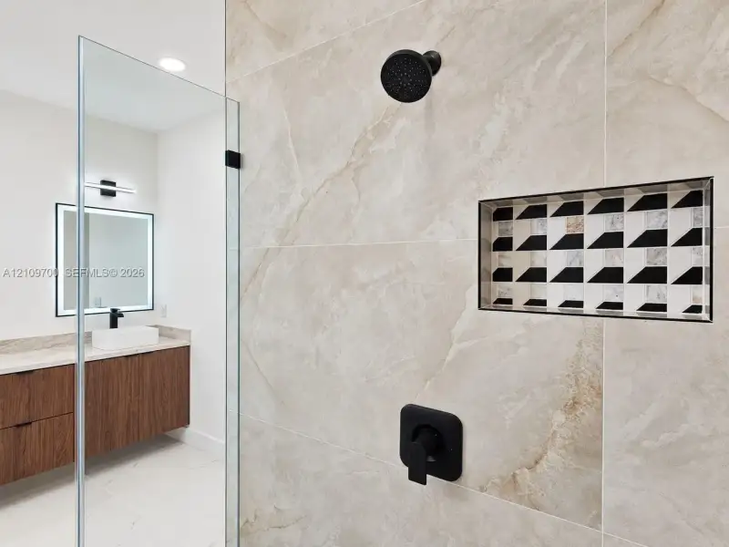 Large-format stone-style tile shower featuring a geometric-patterned recessed niche, matte black rainfall showerhead, and matching control handle, with a wooden-finish floating vanity and vessel sink visible
