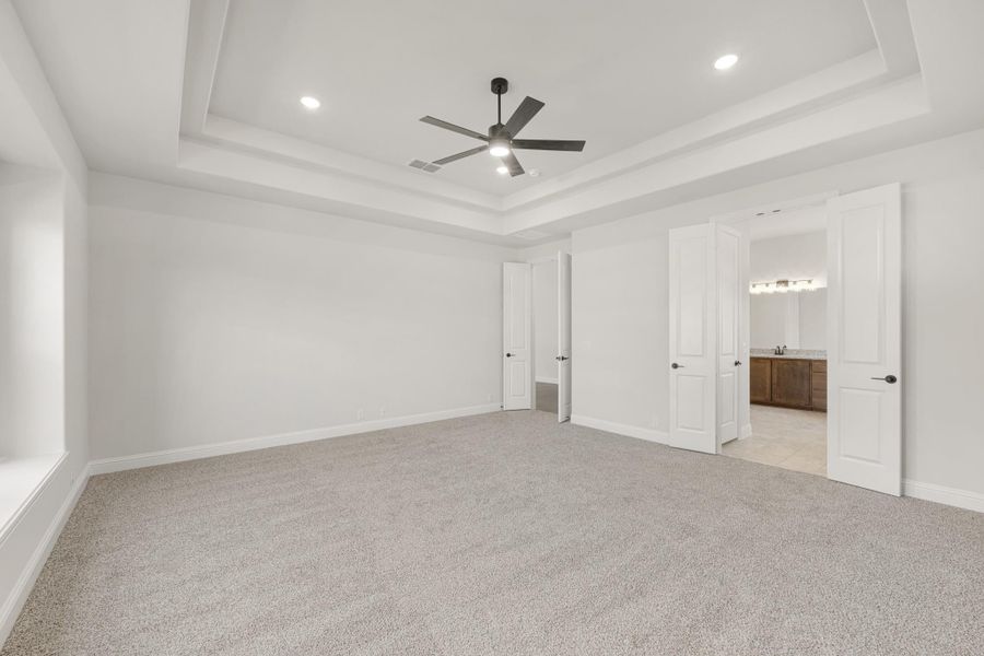 Spacious bedroom with tray ceiling, ceiling fan, carpet flooring, and open door to ensuite bathroom Spacious bedroom with tray ceiling, ceiling fan, carpet flooring, and open door to ensuite bathroom