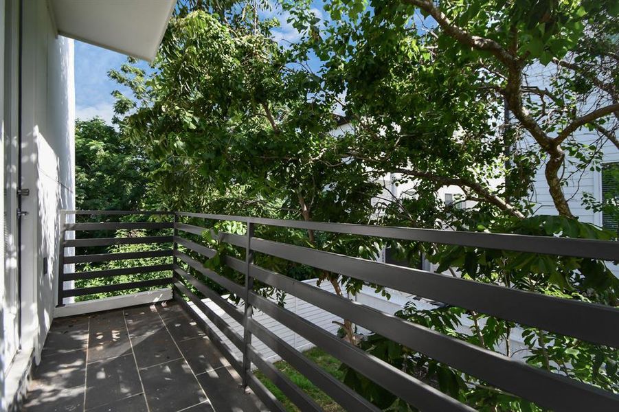 This is a shaded balcony with tiled flooring, metal railings, and a view of lush greenery, offering a peaceful outdoor space adjacent to the home.