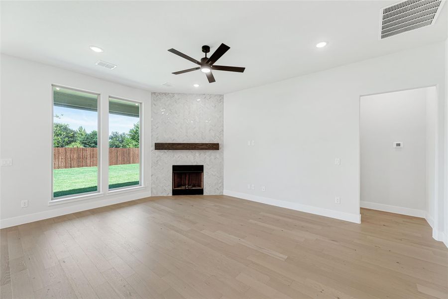 Open living room with light hardwood floors, tiled fireplace, ceiling fan, and large windows overlooking backyard