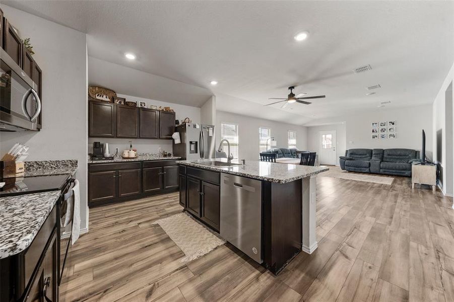 Kitchen featuring stainless steel appliances, dark brown cabinetry, an island with sink, open floor plan, and light wood-type flooring