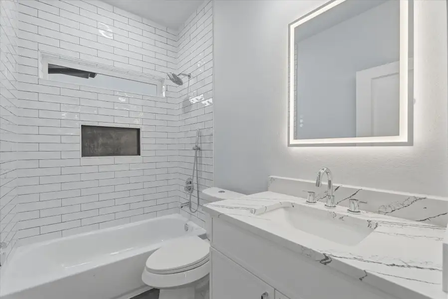 The upstairs full bathroom features classic white subway tile, a shower and tub combination with both standard and handheld shower heads, a modern backlit mirror, and black flooring for a contemporary contrast.
