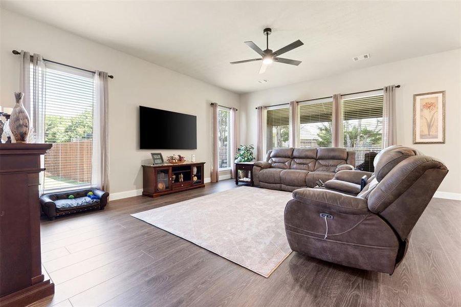 Spacious living area featuring wood-finish flooring, light neutral wall tones, and multiple large windows with blinds Spacious living area featuring wood-finish flooring, light neutral wall tones, and multiple large windows with blinds