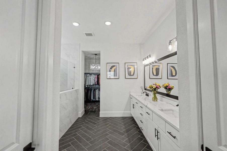 This expansive primary bathroom showcases a white double vanity with quartz countertops and contrasting black hardware. Elegant dark herringbone tile floors lead toward a walk-in shower and a large closet featuring a crystal chandelier.
