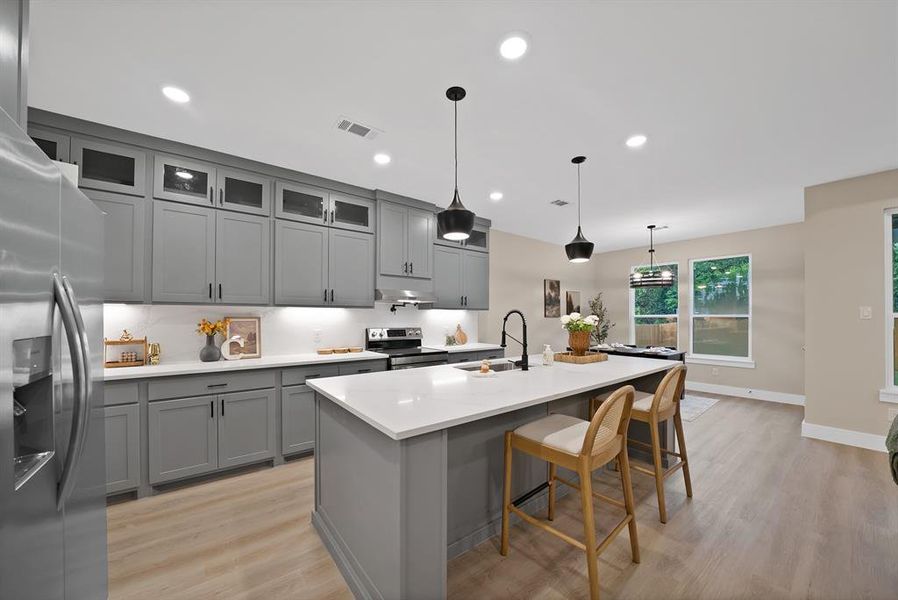 Modern kitchen featuring gray shaker cabinetry, a white countertop island with pendant lighting, integrated stainless steel appliances, and wood-finish flooring Modern kitchen featuring gray shaker cabinetry, a white countertop island with pendant lighting, integrated stainless steel appliances, and wood-finish flooring
