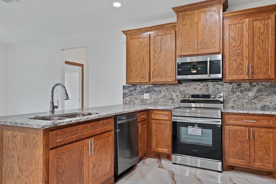 Kitchen featuring brown cabinets, backsplash, light stone countertops, stainless steel appliances, and a peninsula