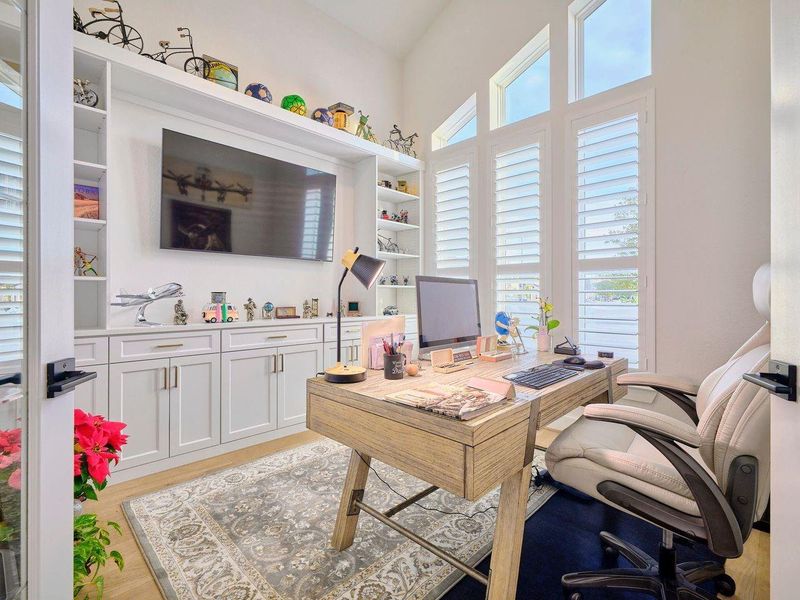 Experience a bright, modern home office with large windows, designer shutters, wide plank flooring, and custom built-in shelving.