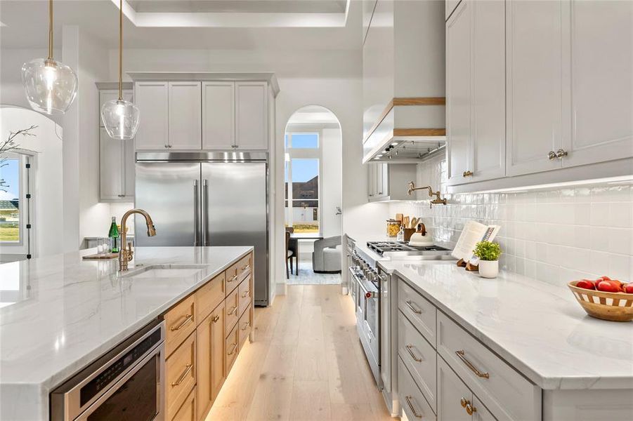 Gourmet kitchen featuring a stainless steel French door refrigerator, a central island with an undermount sink and built-in microwave, light wood-finish flooring, white perimeter cabinetry, and light-toned countertops