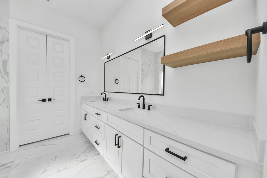 This modern bathroom features a sleek double vanity with white countertops, black fixtures, and ample storage. It includes a large mirror, minimalist lighting, and open wooden shelves for added style. The space is bright and airy, complemented by marble-patterned floors and walls.