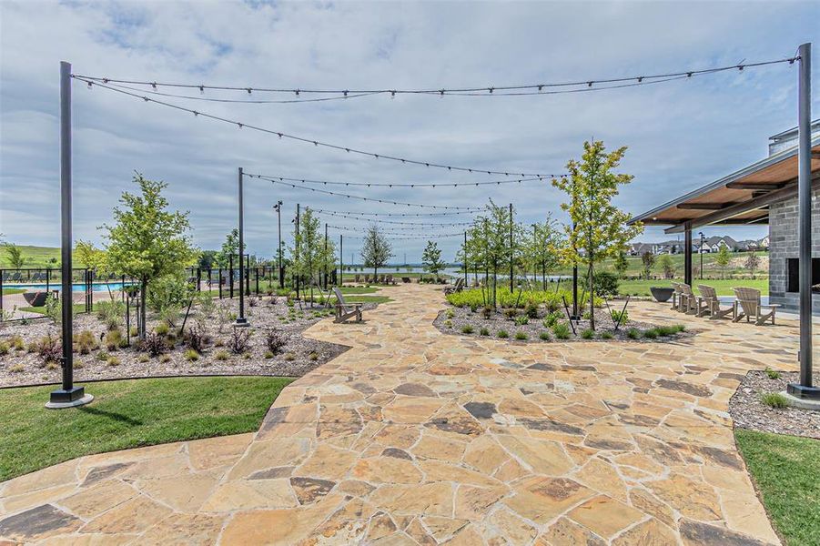 Expansive natural stone patio featuring string lighting, mature landscaping, a shaded patio area with an outdoor fireplace, and a large inground pool