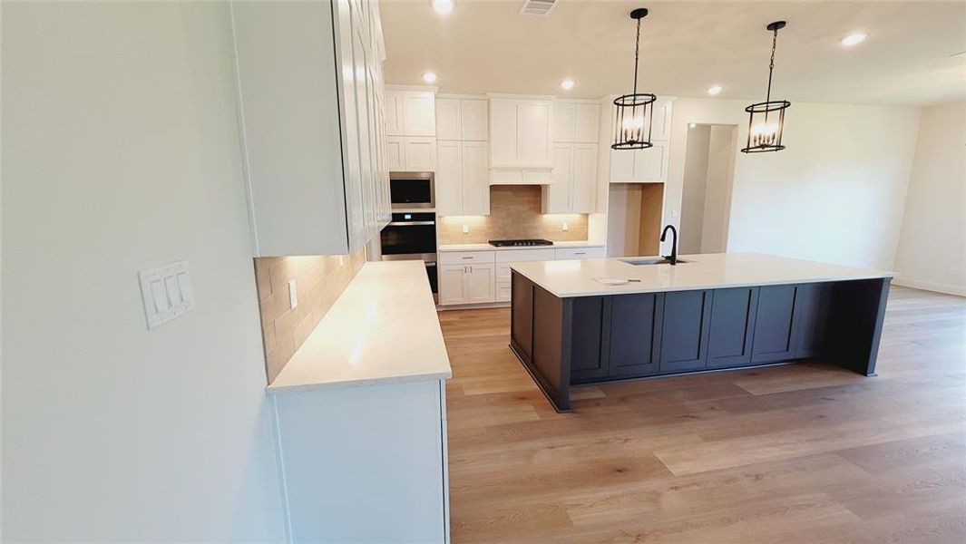 Gourmet kitchen featuring a large central island with a matte black faucet, light wood-finish flooring, white cabinetry, integrated stainless steel appliances, and under-cabinet lighting