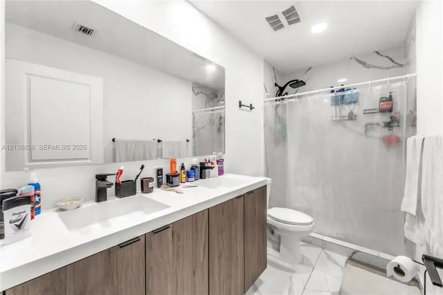 Double vanity featuring wood-finish cabinetry, rectangular under-mount sinks, matte black faucets, and an expansive wall-to-wall mirror, complemented by tiled flooring and a tiled shower surround