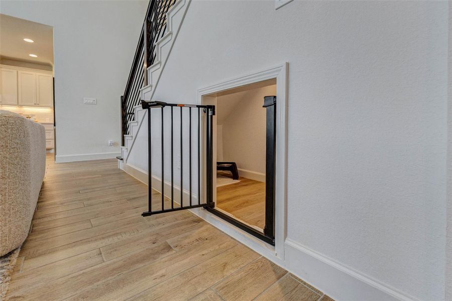 Cozy nook under a staircase, featuring a small door with a safety gate. Ideal for storage or a pet area, it adds functional use to the space. The area is adjacent to an open kitchen with modern features and hardwood flooring. Cozy nook under a staircase, featuring a small door with a safety gate. Ideal for storage or a pet area, it adds functional use to the space. The area is adjacent to an open kitchen with modern features and hardwood flooring.