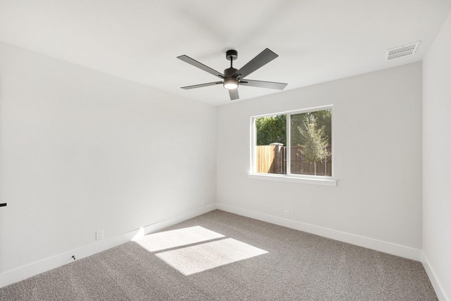 Empty bedroom with gray carpet, white walls, ceiling fan, and a window overlooking a fenced backyard