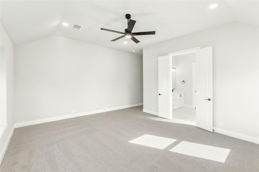 Empty bedroom with gray carpet, vaulted ceiling, ceiling fan, and open door to ensuite bathroom Empty bedroom with gray carpet, vaulted ceiling, ceiling fan, and open door to ensuite bathroom
