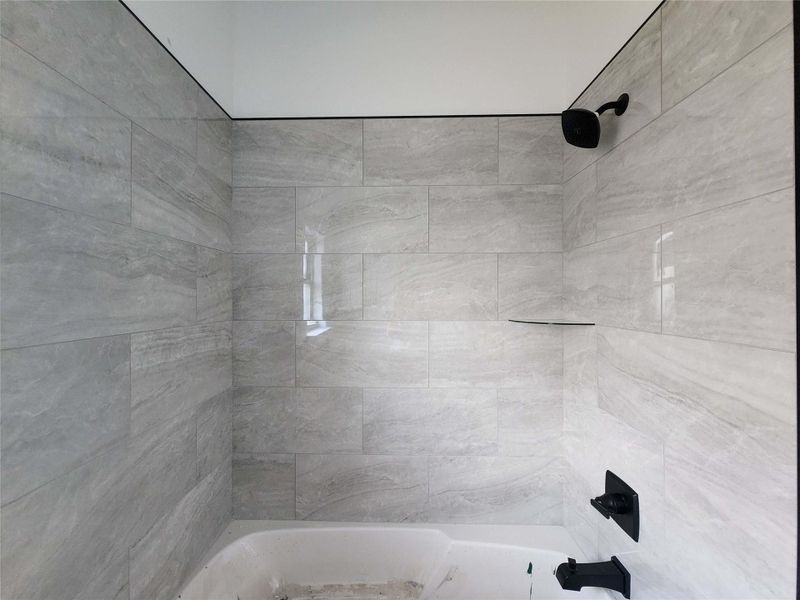 Bathtub with light gray large format tile surround, matte black shower head, matte black faucet, and an integrated corner shelf