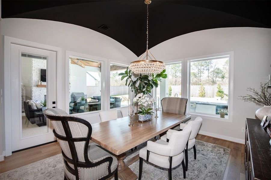 This dining area features a stylish wooden table with elegant chairs, under a striking modern chandelier. Large windows provide abundant natural light and views of the backyard with a pool. The space is complemented by a black arched ceiling and hardwood floors, creating a sophisticated and inviting atmosphere.