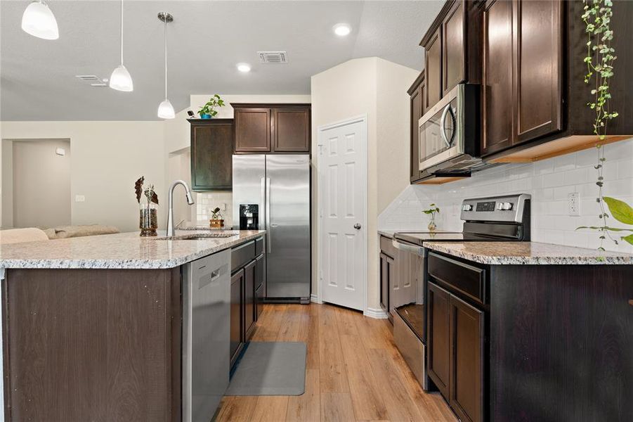 Kitchen featuring stainless steel appliances, dark brown cabinetry, backsplash, light stone counters, and decorative light fixtures