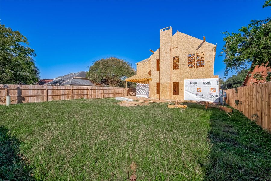 This photo shows a partially constructed house with a wooden frame and some areas covered in Tyvek wrap. It is set in a spacious backyard with a wooden fence, surrounded by greenery.
