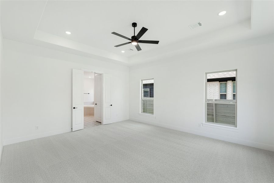 Spacious bedroom with tray ceiling, black ceiling fan, carpet flooring, and open door to ensuite bathroom