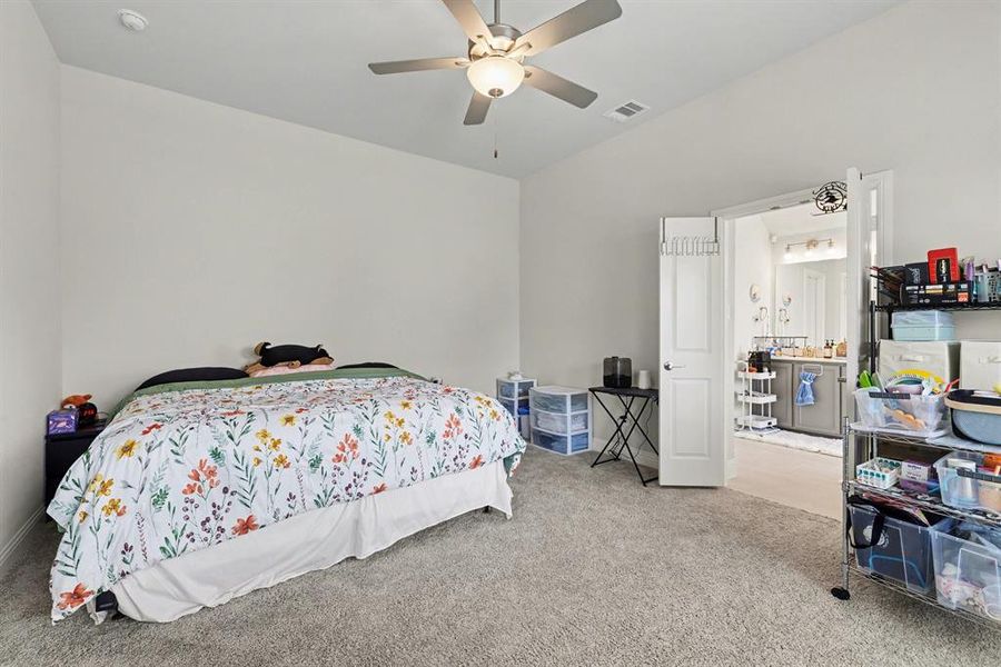 Carpeted room with a ceiling fan with integrated lighting, offering direct access to a bathroom featuring a dual vanity with a light-colored countertop and a wall-mounted light fixture